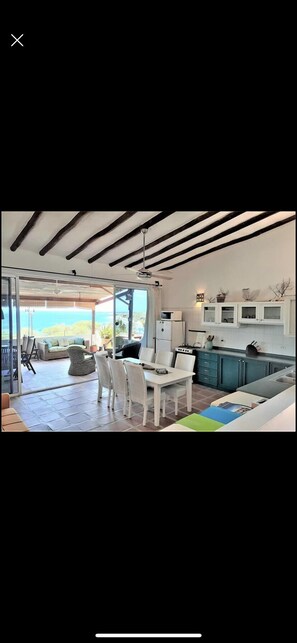 Een moderne keuken met witte kasten, een eethoek met witte stoelen en uitzicht op de oceaan door grote glazen deuren.