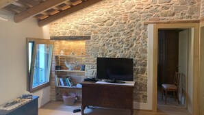 Una camera accogliente con parete in pietra, travi in legno, un televisore su un mobile in legno, una libreria e una finestra.