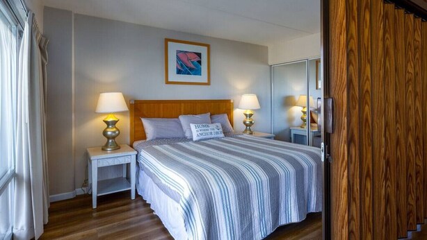 A hotel room with a bed, two bedside tables with lamps, a wooden headboard, a framed picture on the wall, and a wooden door. - 1519 · Ilikai Collection # 9. Beachfront Resort, upgraded (Honolulu)