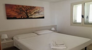 A bedroom with a bed, a nightstand, a lamp, a window with blinds, and a painting on the wall. - Two-room apartment with ocean view on the 2nd floor - Afrodite (Torre Mozza)