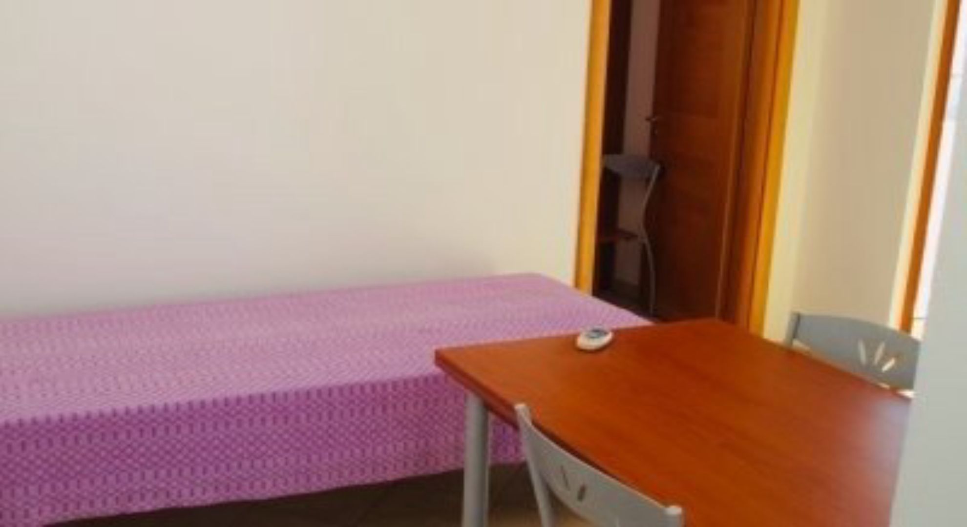 Ein kleines Zimmer mit einem Einzelbett, einem Holztisch und einem Stuhl.