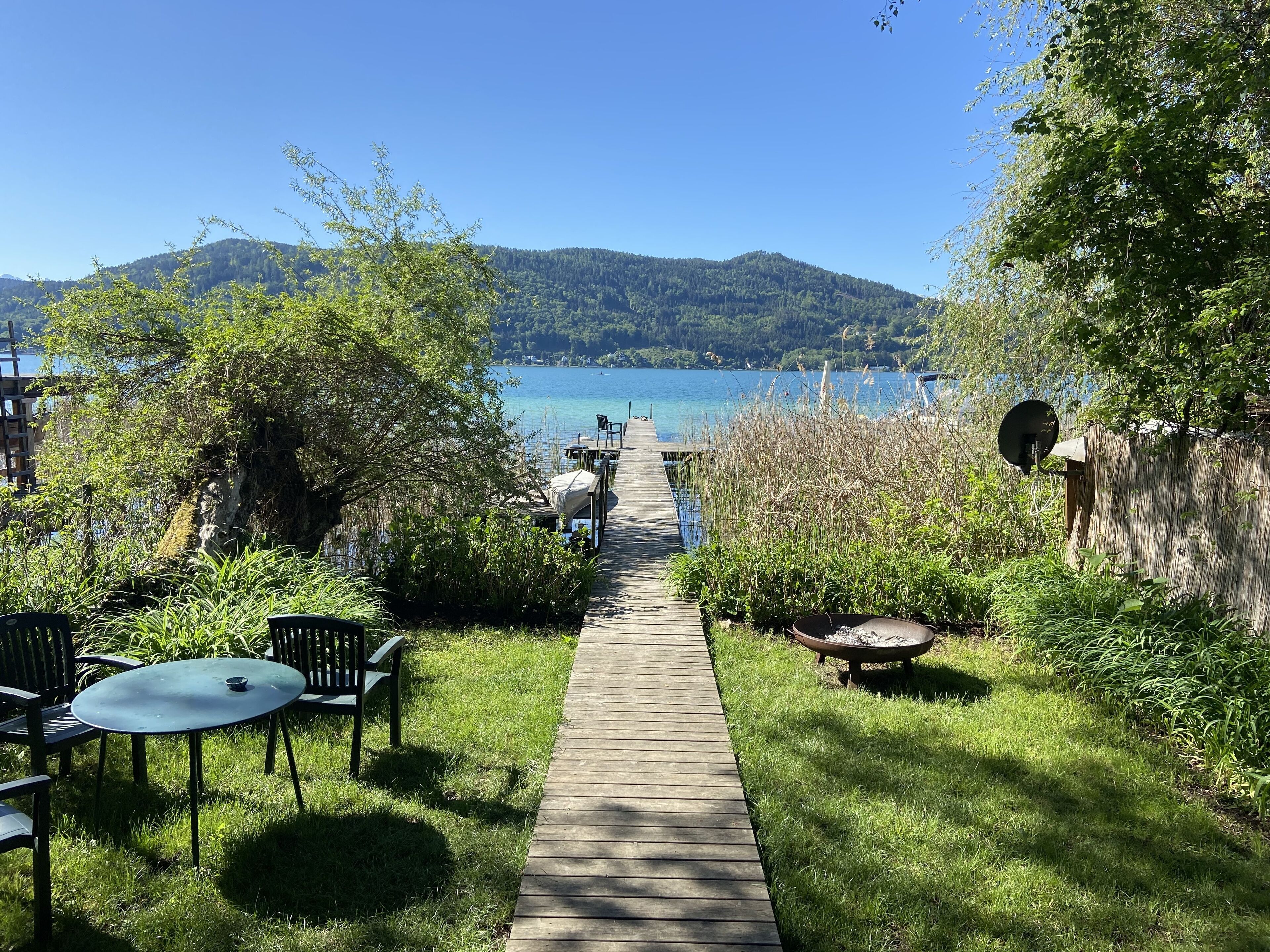 A wooden deck leading to a lakeside area with chairs and a table, surrounded by greenery and a view of the lake and hills.