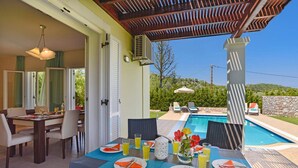 A dining area with a table set for four, a poolside lounge with chairs, and a view of a swimming pool. - Apollon - Seven Springs Villas (Rhodes)