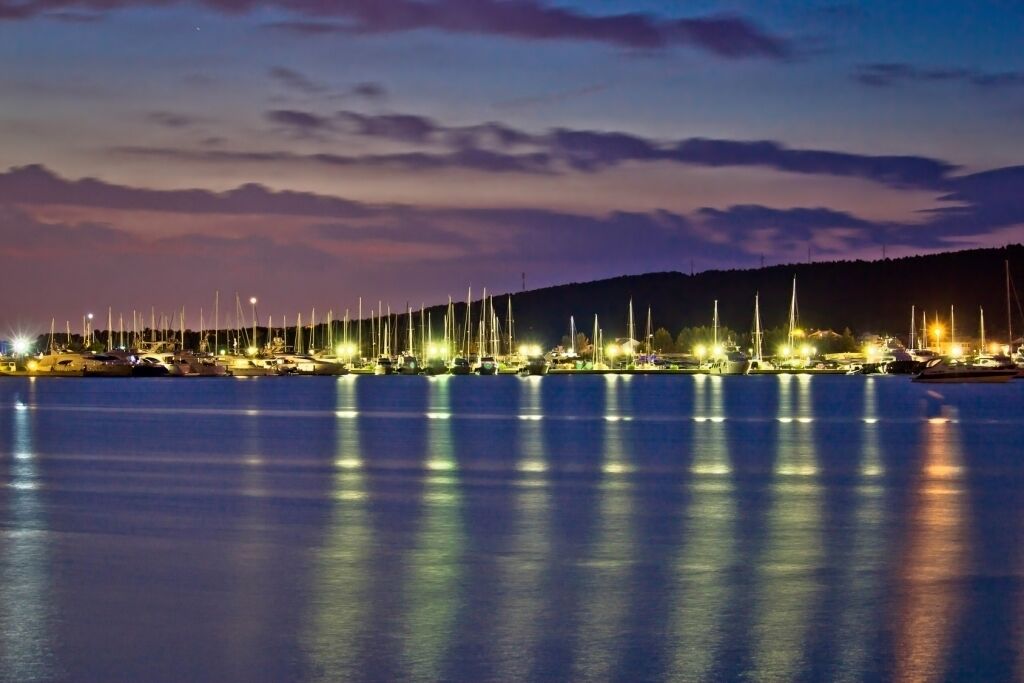 A marina at dusk with numerous boats docked and illuminated by lights.