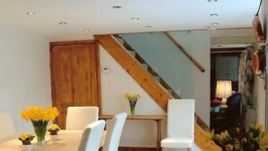 Un coin repas avec une table en bois et des chaises blanches, une petite table d’appoint agrémentée de fleurs et un escalier avec des rampes en bois.