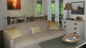Un salon avec un canapé beige, deux coussins, une table basse, une lampe, une table d’appoint avec des livres et un tableau au mur.