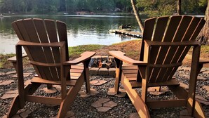 Zwei Adirondack-Stühle mit Blick auf einen See, mit einem Steg und einem Feuerkorb im Vordergrund.