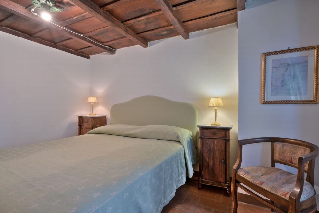 Um quarto com uma cama, duas mesas de cabeceira com luminárias, uma cadeira de madeira e um quadro na parede.