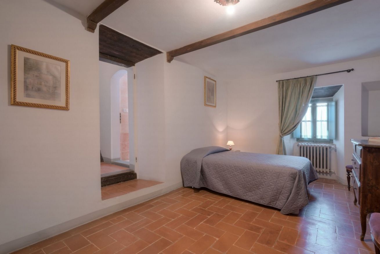Ein Schlafzimmer mit Bett, Heizkörper, einem Fenster mit Vorhängen, einem Holzbalken an der Decke und einem gerahmten Bild an der Wand.