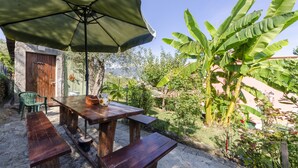 An outdoor wooden table with benches and a green umbrella, surrounded by lush greenery and a stone building. - Casa Rosa, Rapallo (rapallo)