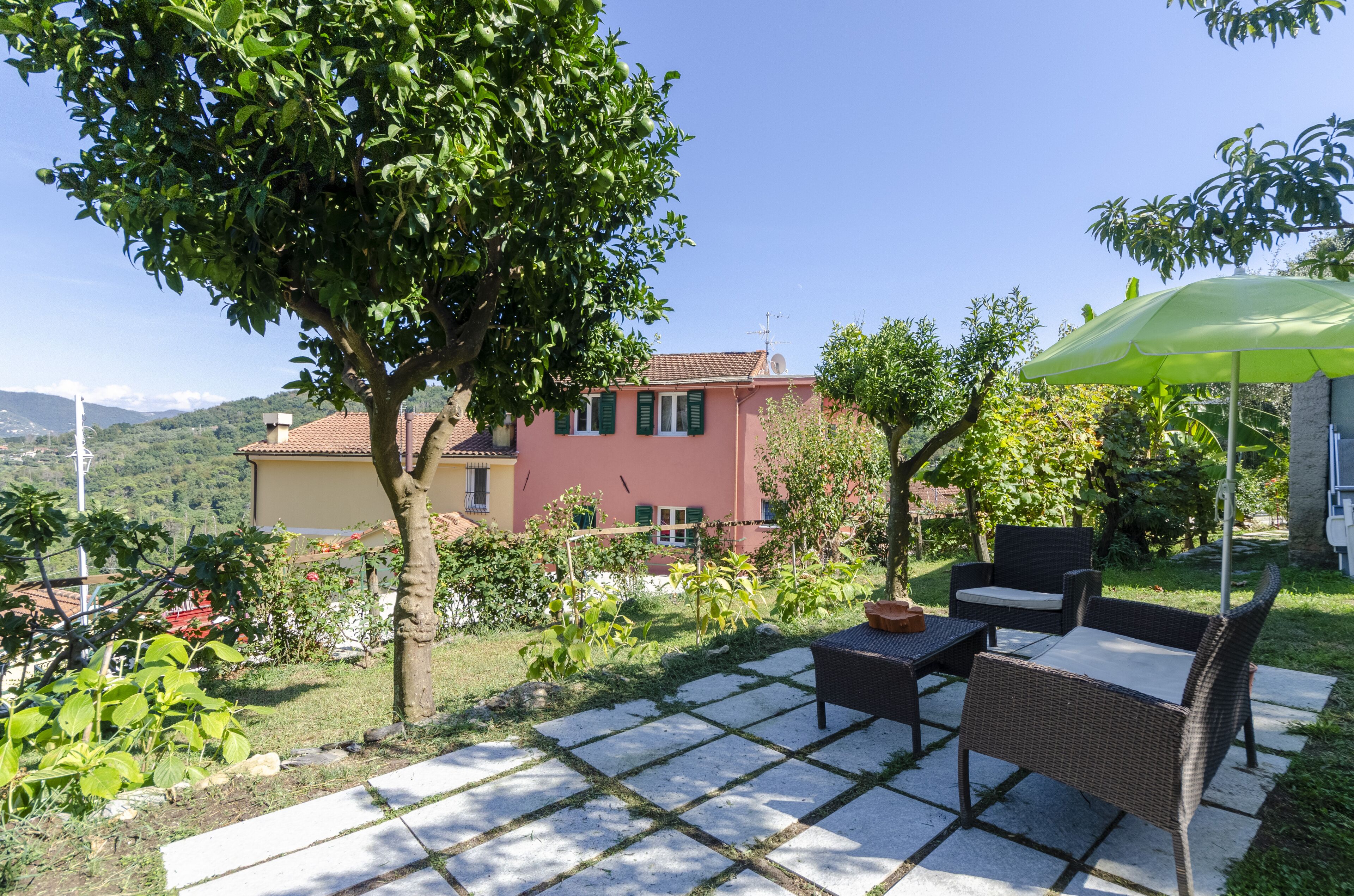 Une terrasse avec des meubles d’extérieur, un parasol vert et une vue sur une maison rose dotée d’un jardin.