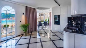 A modern hotel room with a large window offering a view of the beach, a dining area with chairs and a table, and a kitchen area with a counter and cabinets. - Crown of La Concha Beach by DonosRent (San Sebastián)