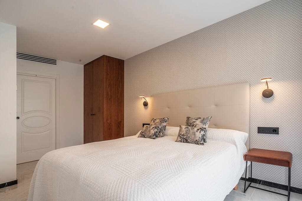 A bedroom with a large bed, two wall-mounted lights, a wooden wardrobe, and a bench.