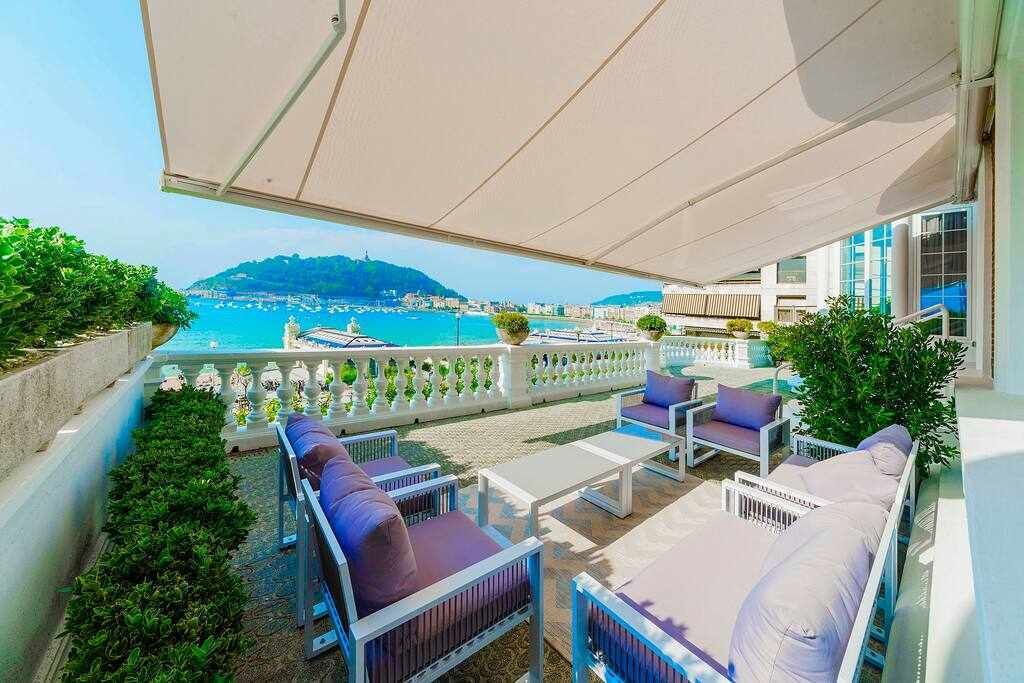 A balcony with outdoor seating, a view of the sea, and a mountain in the distance.