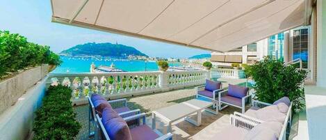 A balcony with outdoor seating, a view of the sea, and a mountain in the distance.