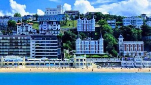 A coastal cityscape with a variety of buildings, including a prominent white structure with a pointed roof, a modern building with a glass facade, and several residential buildings. - Crown of La Concha Beach by DonosRent (San Sebastián)