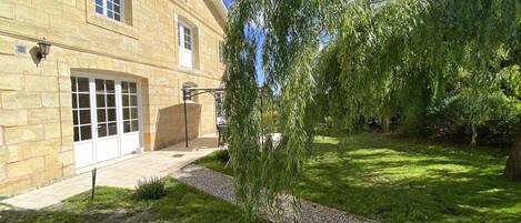 A stone building with large windows and a garden with a weeping willow tree.