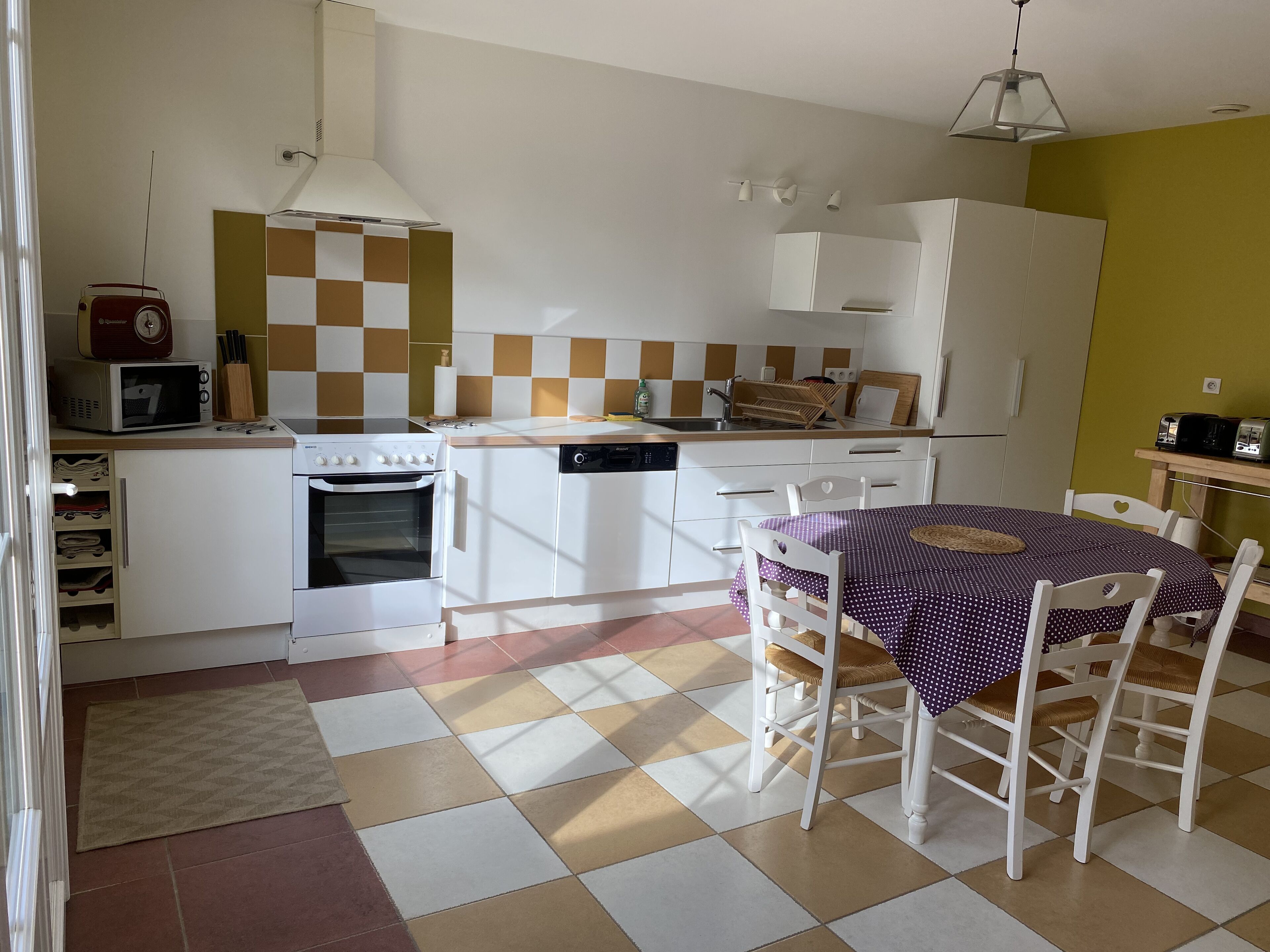A compact kitchen with a microwave, stove, and dishwasher. It features a checkered floor, a dining table with chairs, and a hanging light fixture.