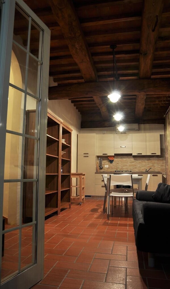 A rustic kitchen with wooden beams, a dining area with a table and chairs, and a sliding glass door leading to a storage area.