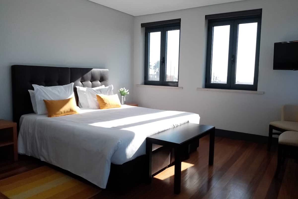 A bedroom with a large bed, two windows, a TV, and a wooden floor.