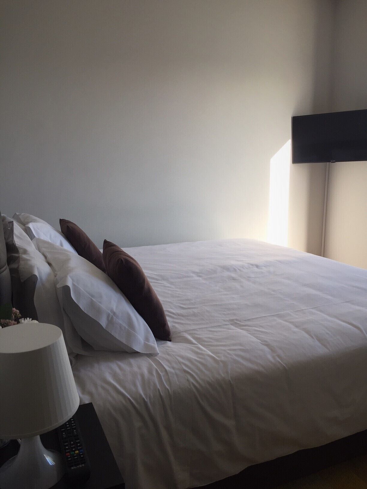 A neatly made bed with white and brown pillows, a remote control, and a lamp on a bedside table.
