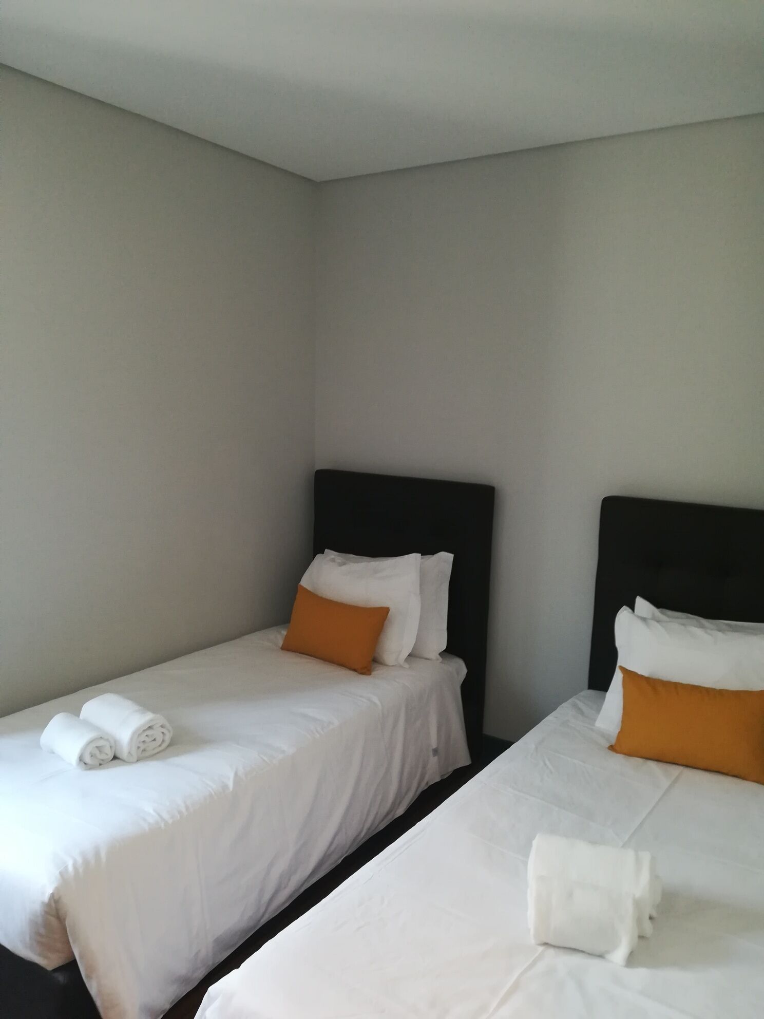 A hotel room with two single beds, each with white bedding and orange pillows, and a neatly made white towel on the floor.