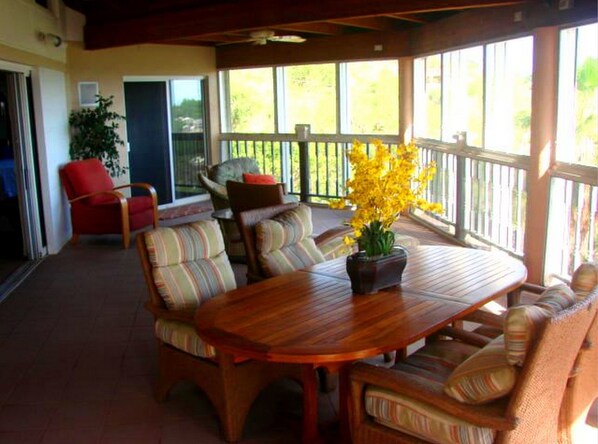 Un patio cubierto con una mesa de madera y sillas con cojines a rayas, un sillón rojo y una planta en maceta con flores amarillas.