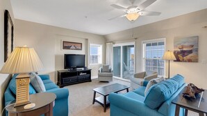 Ein Wohnzimmer mit Deckenventilator, einem Fernseher auf einer schwarzen Unterhaltungseinschub und blauen Sofas, einem Couchtisch und einem Bild an der Wand.