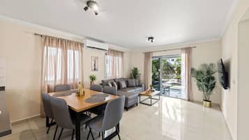A modern living room with a dining area, a sofa, a TV mounted on the wall, and a view of the outdoors.