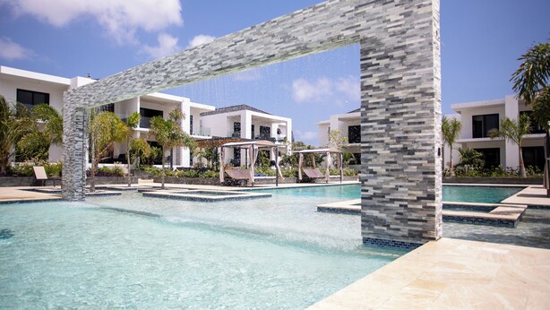 Una piscina con un arco in pietra, sedie a sdraio e moderni edifici sullo sfondo.