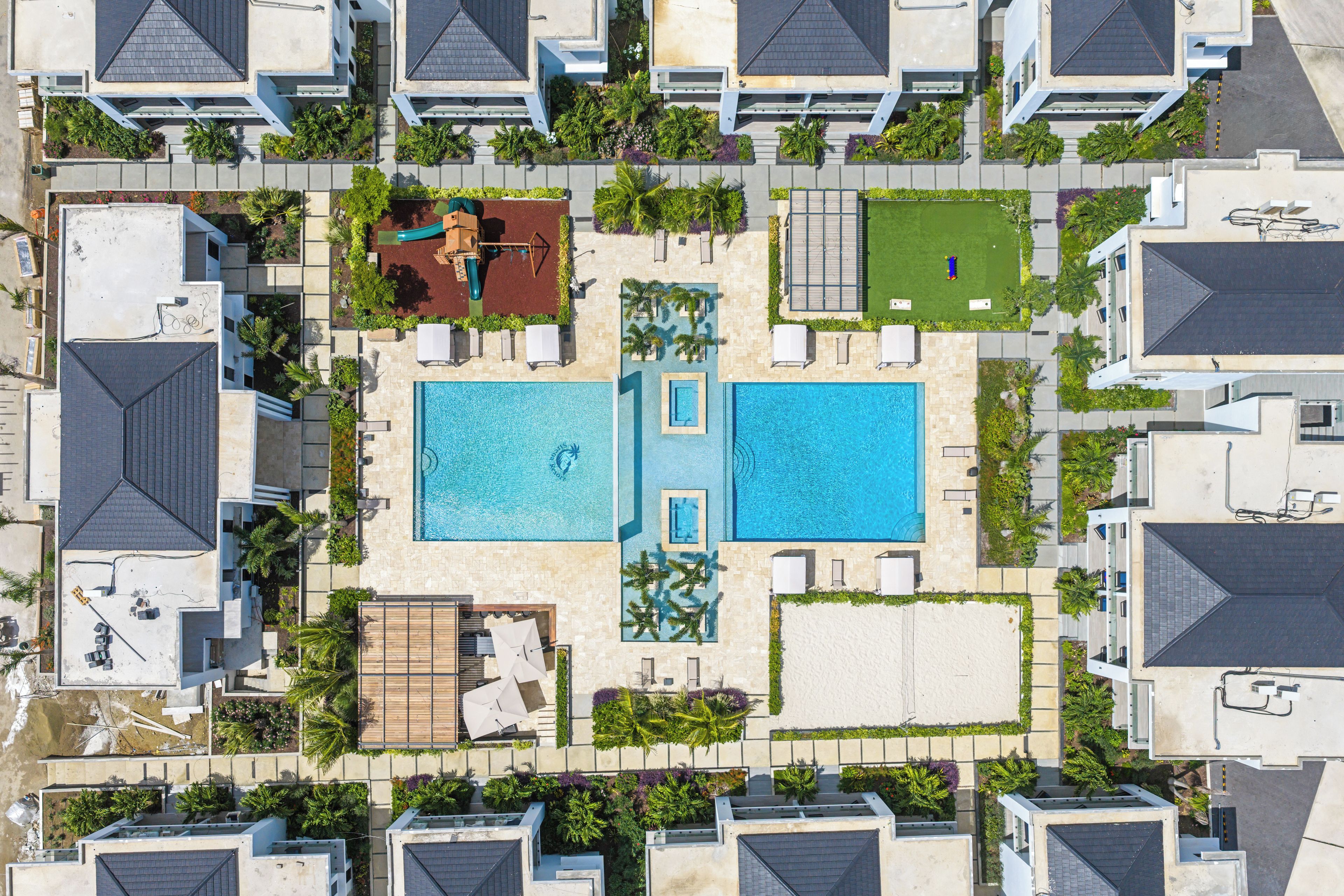 Vista aérea de un complejo turístico con dos piscinas, una zona de juegos para niños y una cancha de tenis.