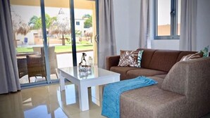 Una sala de estar con un sofá marrón, una mesa de centro blanca y un ventanal que da a un área exterior con patio y vistas de un edificio.