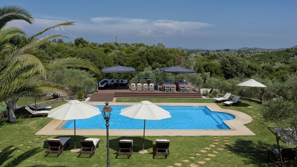 Une piscine bordée de chaises longues et d’ombrelles, entourée d’une végétation luxuriante et avec des collines en arrière-plan.