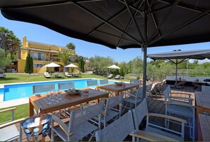 Un espace extérieur avec une piscine, des tables et des chaises en bois, des parasols, et une grande maison en arrière-plan.