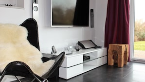 A modern living room with a large flat-screen TV mounted on the wall, a glass coffee table, a white sofa, and a wooden stool.