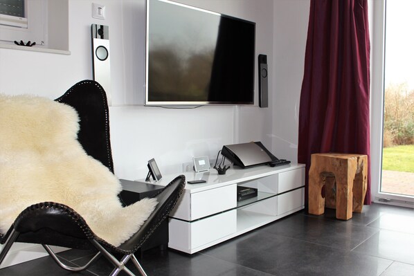 A modern living room with a large flat-screen TV mounted on the wall, a glass coffee table, a white sofa, and a wooden stool.