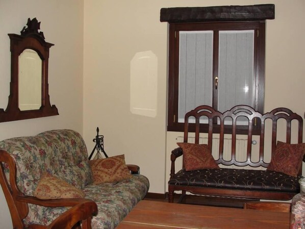 A living room with a floral sofa, a dark upholstered bench, a wooden chair, a mirror, and a window with curtains.