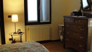 Una camera da letto con un letto, una cassettiera, una lampada, una finestra, un termosifone e un piccolo tavolo.