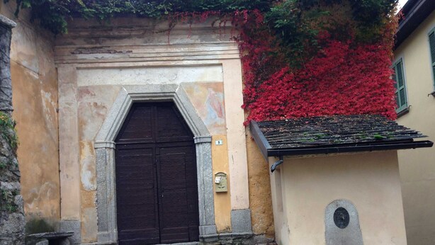 Un edificio in pietra con una porta in legno scuro, un piccolo pozzo in pietra e fogliame rosso sulla parete.