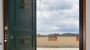 Une porte verte avec une poignée en laiton, qui donne sur une terrasse carrelée offrant une vue sur un paysage vallonné et illuminée de guirlandes lumineuses.