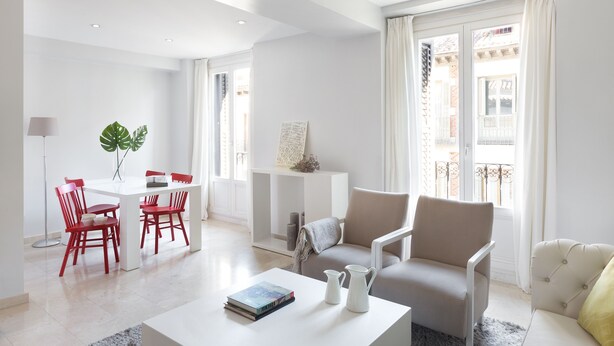 Ein modernes Wohnzimmer mit einem weißen Tisch, roten Stühlen, einem beigen Sofa und einem großen Fenster mit Vorhängen.