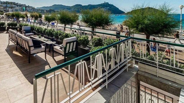 A terrace with outdoor seating, a railing, and a view of the beach and sea.