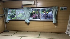 A room with tatami mat flooring, a large window with curtains, and an air conditioning unit on the wall. - Minshuku Teruya (Nago)