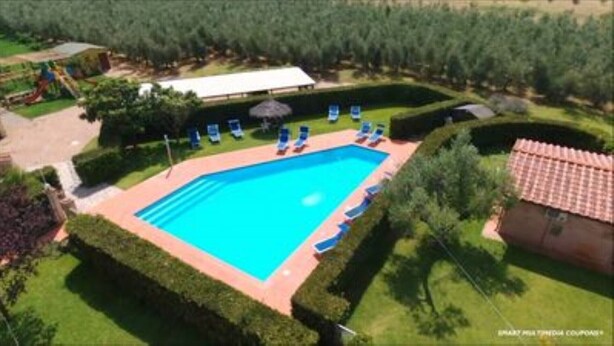 Uma piscina retangular com deck ladrilhado, cercada por sebes bem cuidadas e algumas cadeiras de sol.