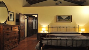 Quarto com cômoda de madeira, cama com cabeceira de metal, duas luminárias de apoio, um quadro na parede e uma porta que dá para outro cômodo.