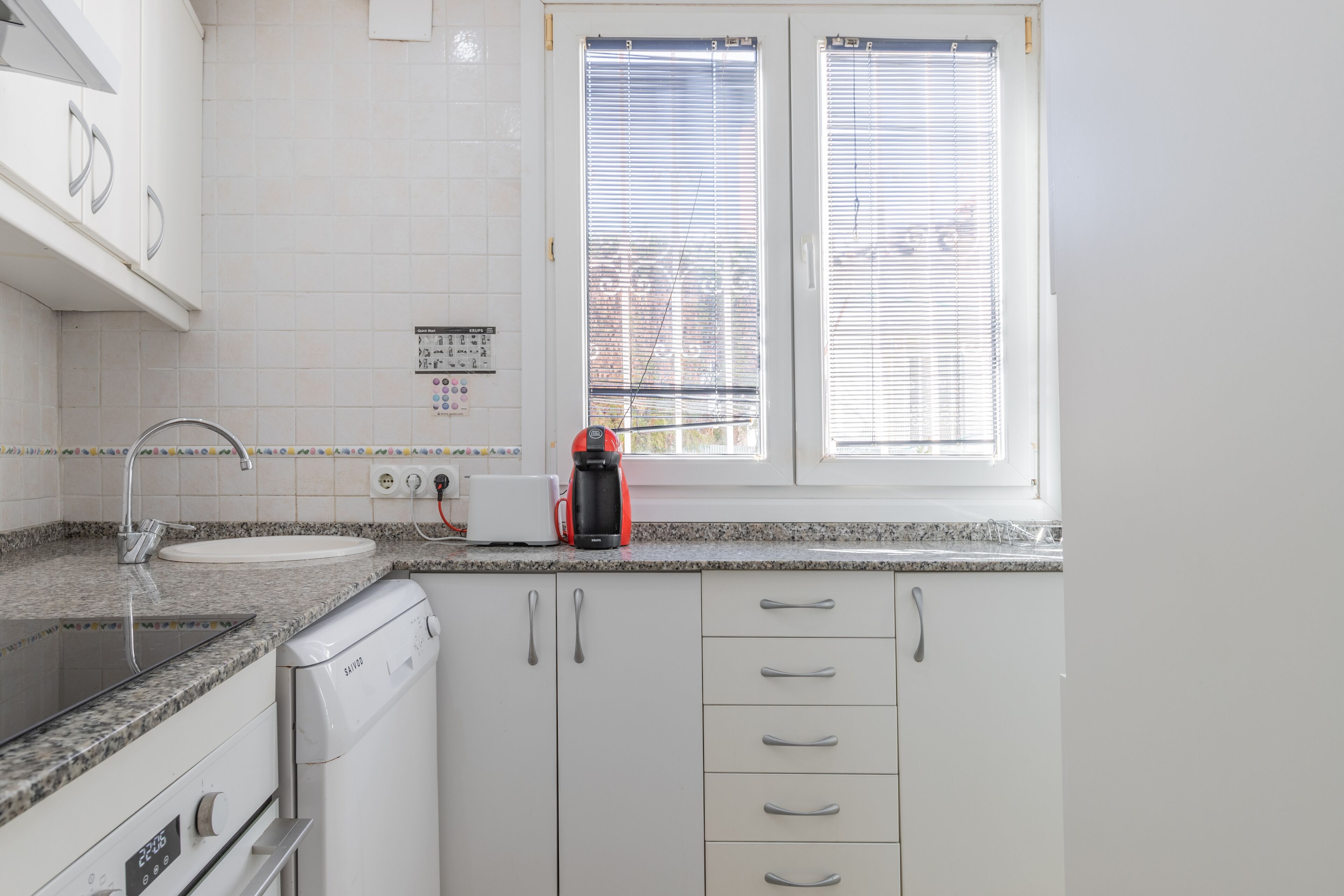 A kitchen with white cabinets, a granite countertop, a coffee maker, and a window with blinds.
