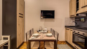 A compact kitchen with wooden cabinets, a dining table set for two, a built-in oven, and a flat-screen TV mounted on the wall.