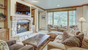 A spacious living room with a stone fireplace, comfortable seating, and large windows offering a view of the outdoors.