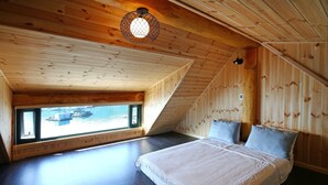 A bedroom with a wooden sloped ceiling, a large bed with white bedding, and a view of boats on water through a window. - Tongyeong Daore Pension (Tongyeong)
