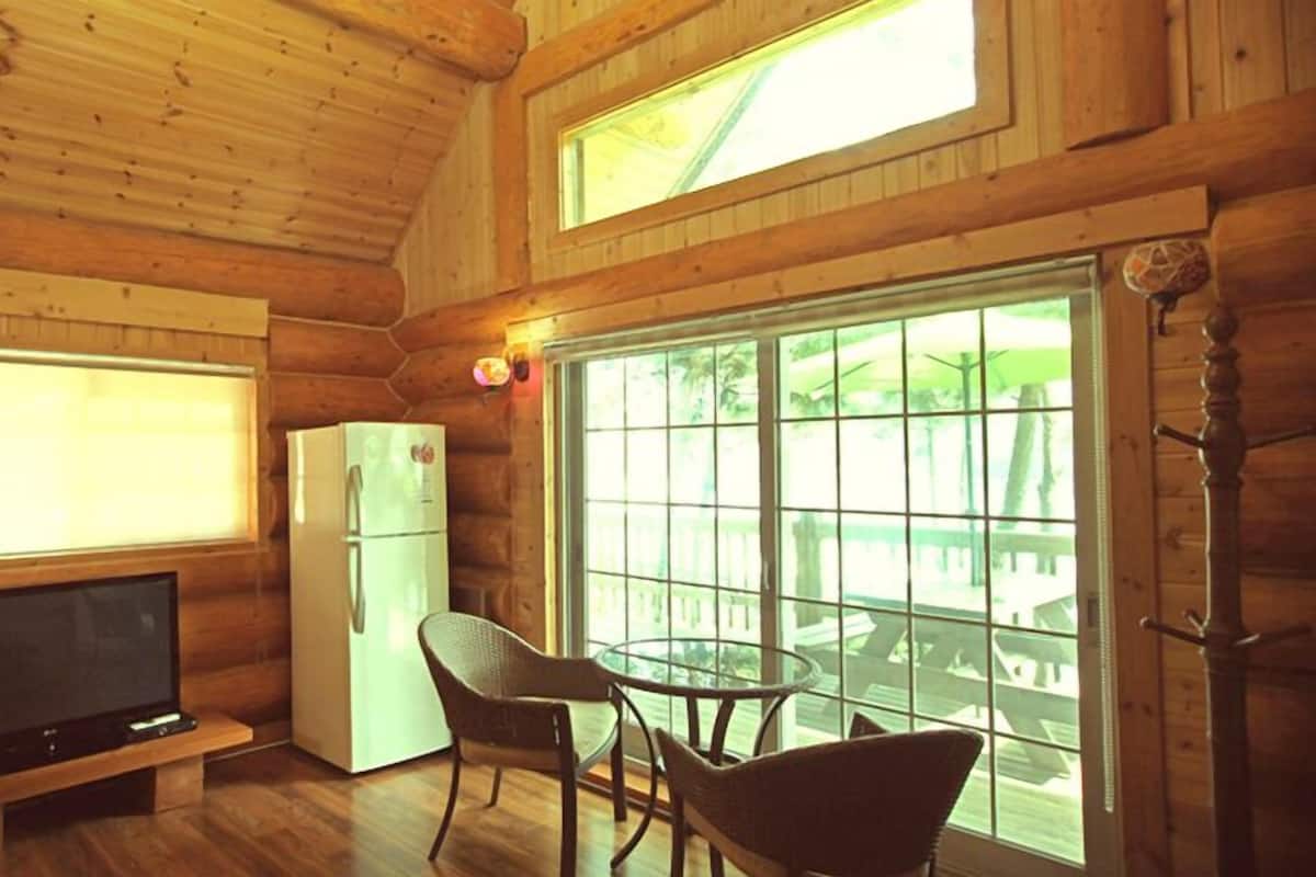 Eine Hütte mit Holzvertäfelung, einem Fernseher auf einem Holztisch, einem Kühlschrank und einem Glastisch mit Stühlen.